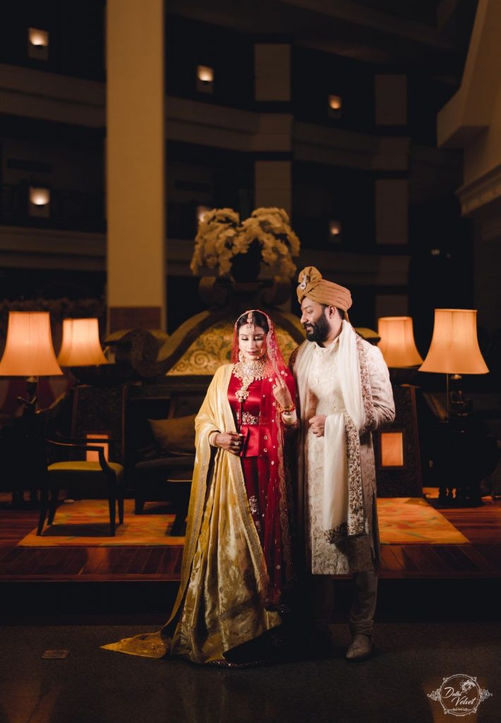 red lehenga with golden dupatta and groom in cream kurta with copper headgear for beach wedding 